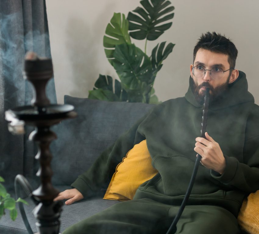 A man with a beard sitting on a couch alone while smoking out of a hookah.